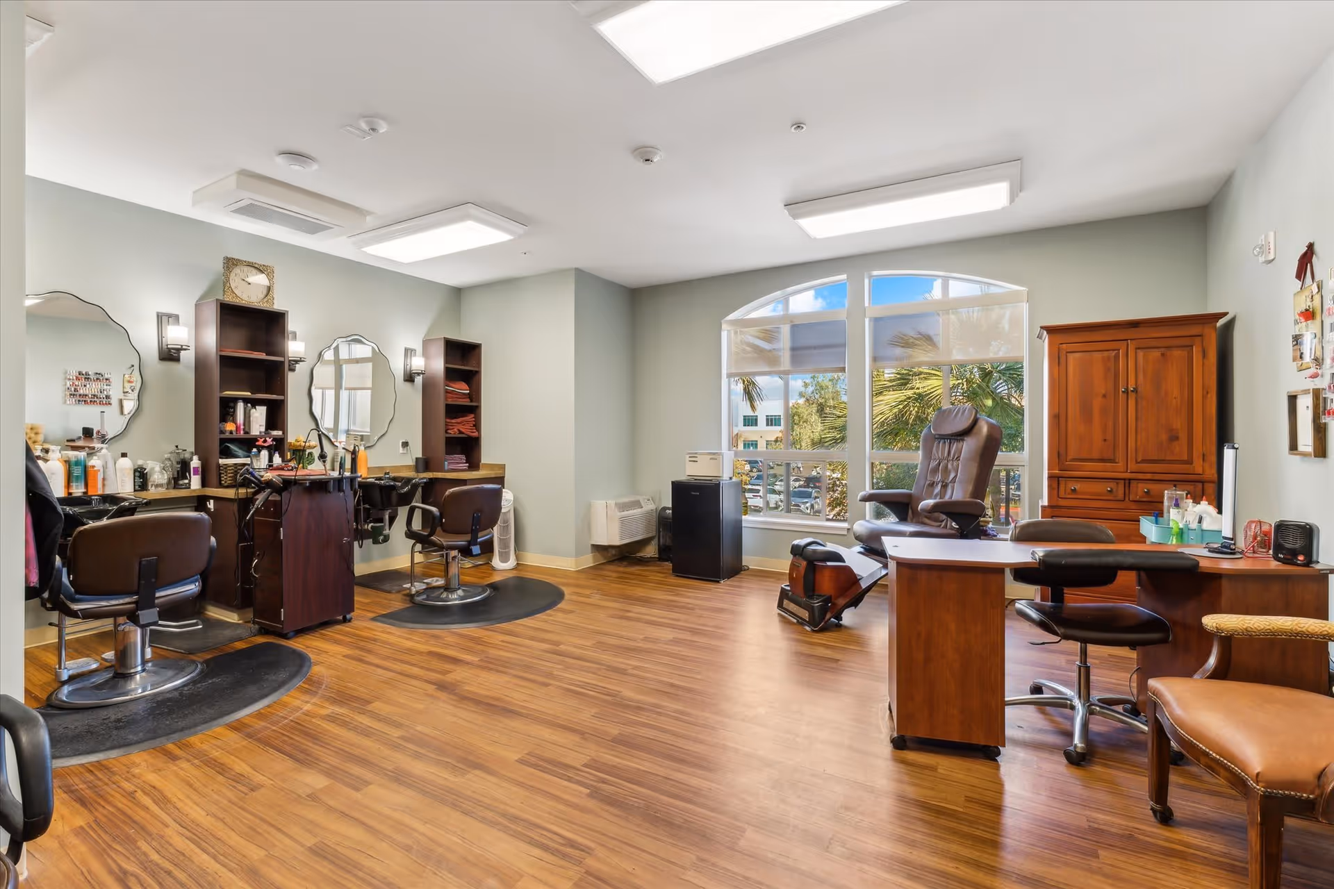 A bright and spacious salon room with wooden flooring, two styling chairs in front of mirrors and shelves stocked with hair products, a pedicure chair near a large window with a view of palm trees outside, and a wooden cabinet and desk with a rolling chair.