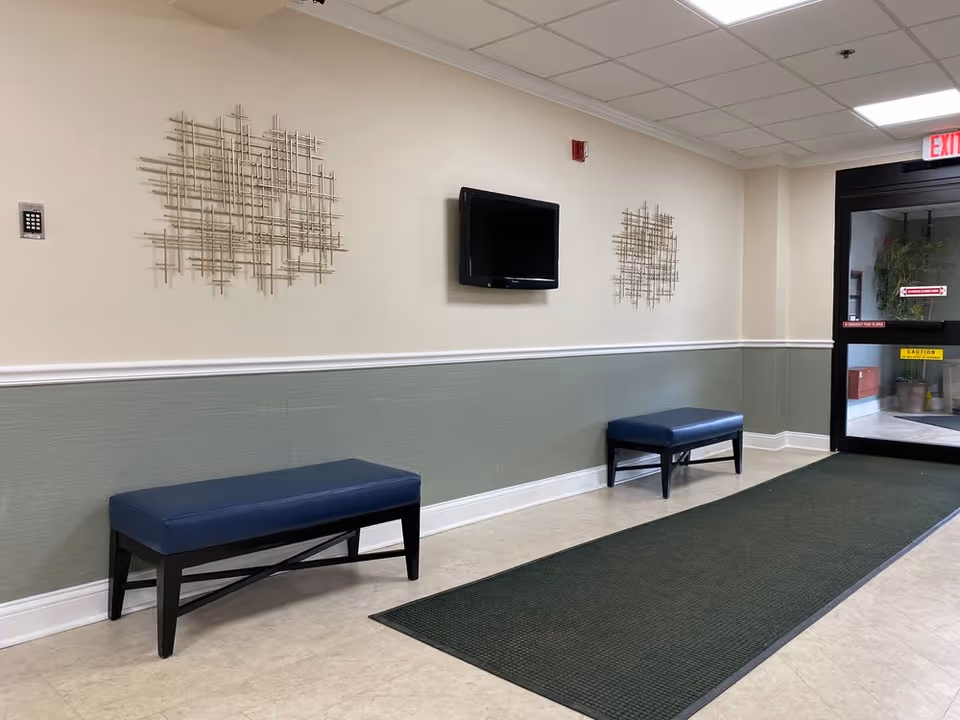Interior hallway of a senior living facility with two blue cushioned benches along the wall, a wall-mounted flat screen TV, decorative metal wall art, and a glass exit door at the end of the hallway.