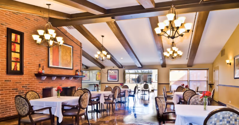 A spacious dining room with multiple tables covered in white tablecloths, each adorned with a small vase of red flowers. The room features a brick wall with a fireplace, wooden ceiling beams, and several chandeliers providing warm lighting. Large windows allow natural light to fill the space.