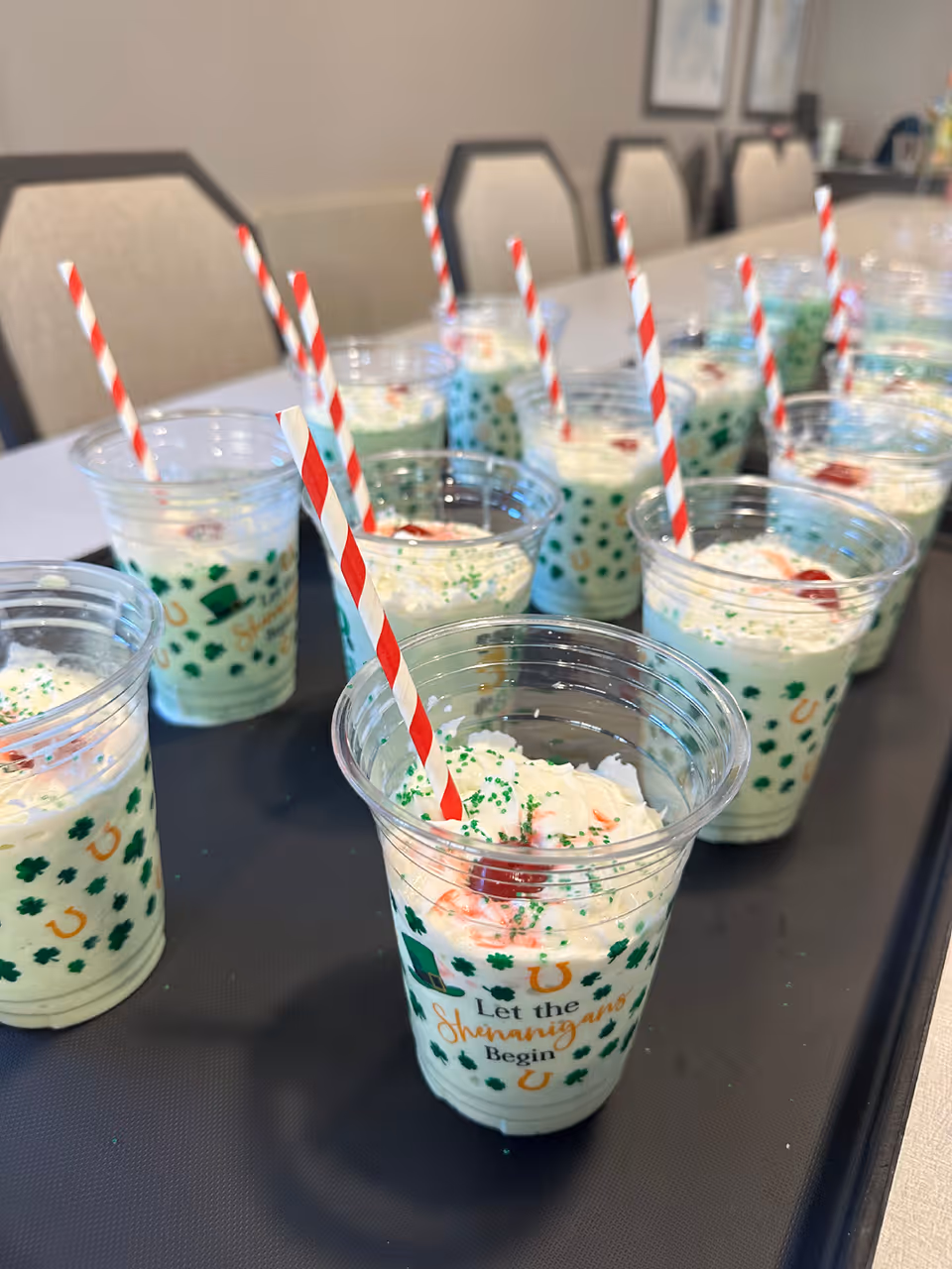 Rows of festive green dessert cups topped with whipped cream, cherries and red-striped straws on a table in a dining area.