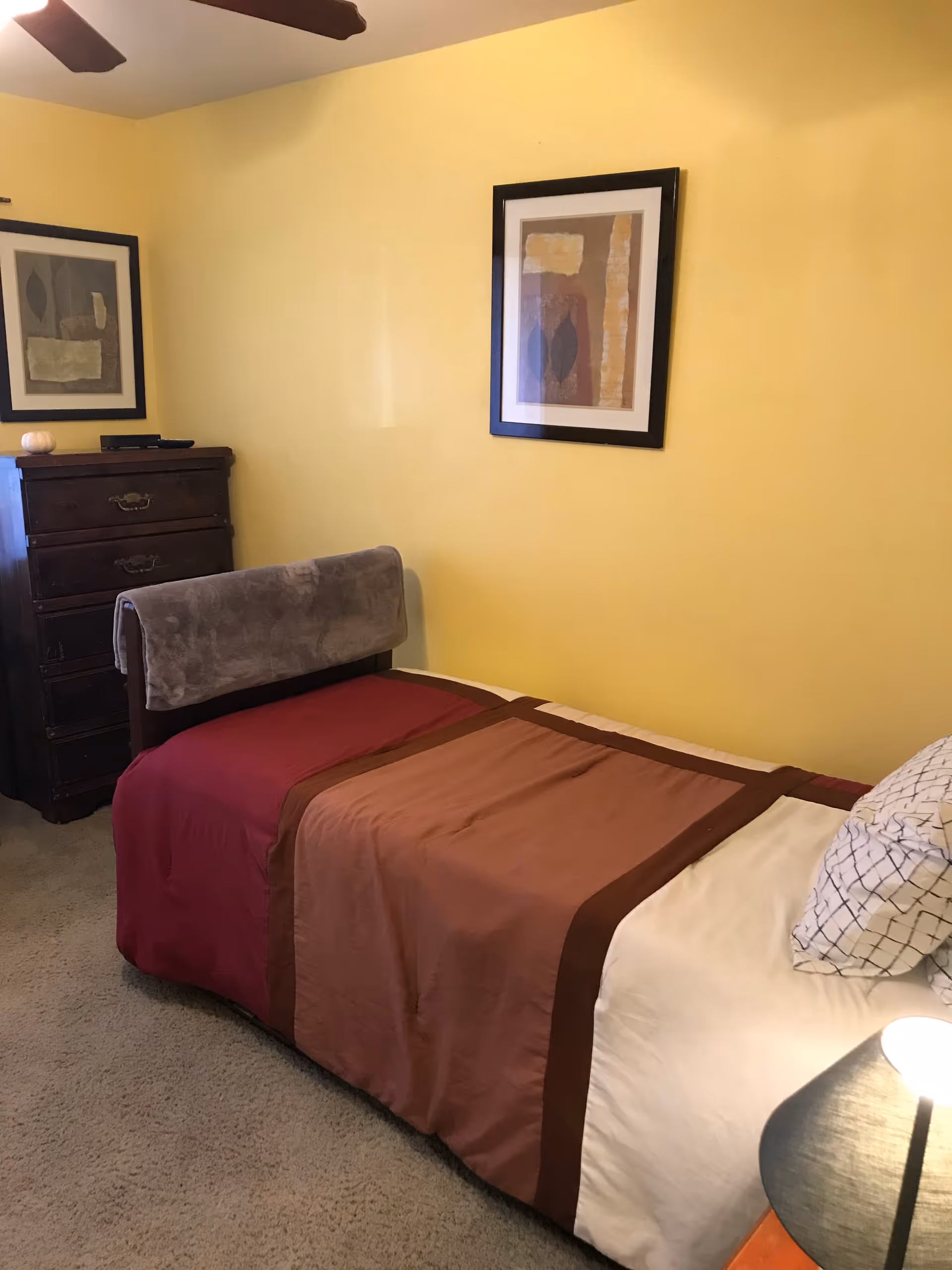 A small bedroom with a single bed covered in a red and brown bedspread, a pillow with a geometric pattern, a wooden dresser with drawers, two framed abstract art pieces on yellow walls, and a ceiling fan above.