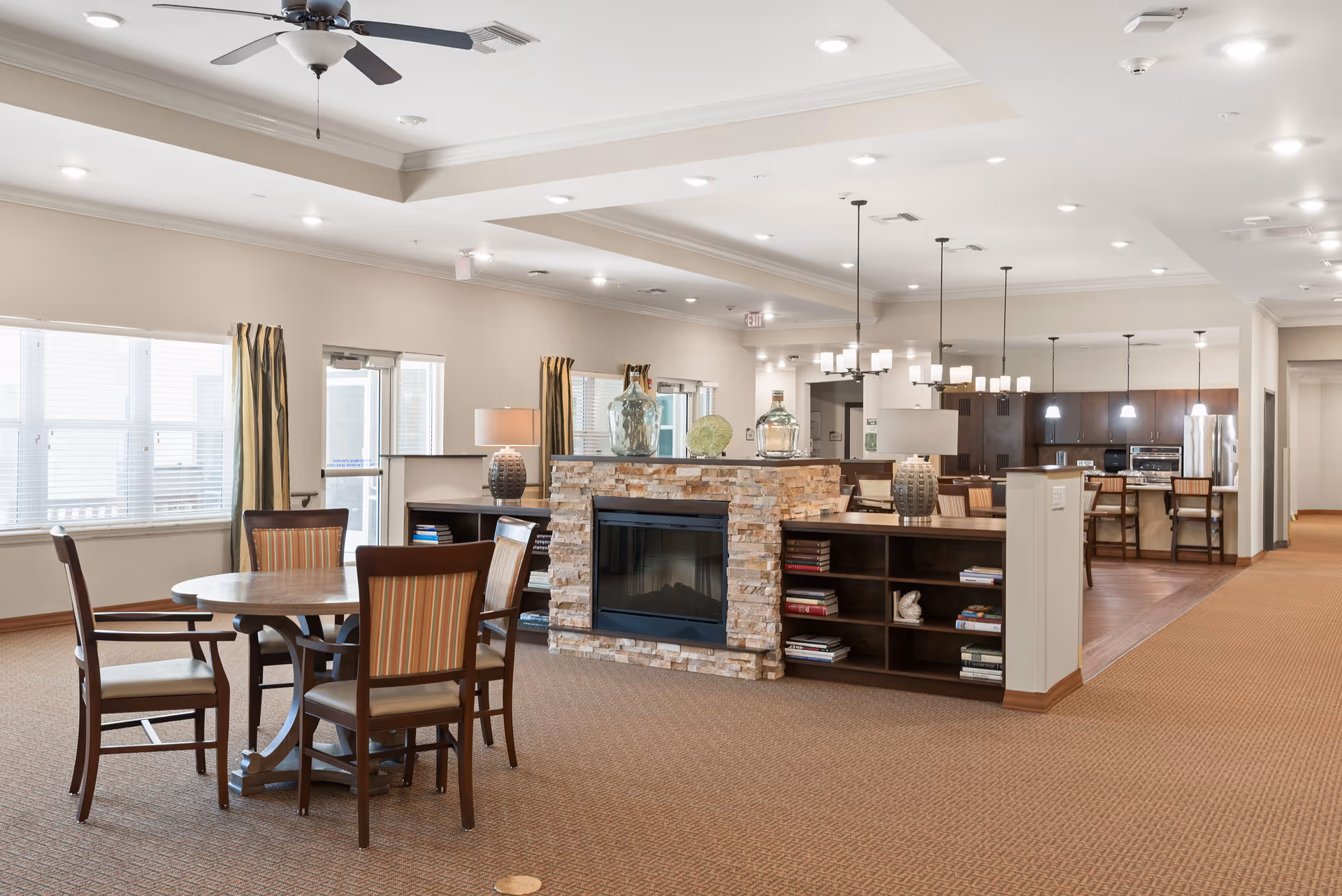 Bright communal lounge and dining area with a round table and chairs, a central stone fireplace and shelving, and an open kitchen in the background.