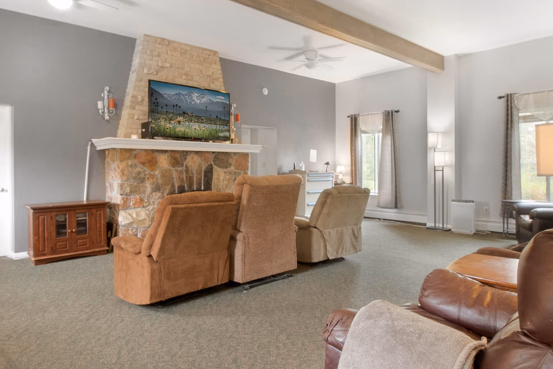 A spacious living room with a stone fireplace and a mounted flat-screen TV displaying a scenic mountain view. The room features several comfortable recliner chairs arranged facing the fireplace and TV. There are windows with curtains allowing natural light to enter, and the walls are painted in a soft gray color. The room has a carpeted floor and is decorated with lamps and a wooden cabinet.
