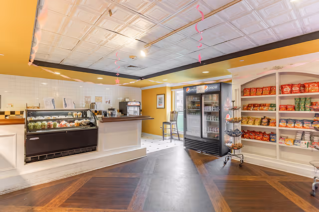 Interior view of a small convenience or snack area in a senior living facility with a refrigerated display case containing food, a Pepsi-branded refrigerator stocked with beverages, shelves filled with various snack bags, and a counter with a coffee machine and other items. The room has a white tiled ceiling, yellow walls, and dark wood flooring with a geometric pattern.