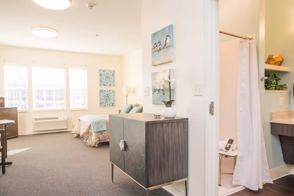 A bright and spacious senior living room with a bed covered in patterned bedding and a light blue throw blanket. The room has three large windows with white blinds, a wooden dresser, and a small cabinet with decorative artwork above it. To the right, there is a bathroom area with a shower curtain partially drawn and a shower chair visible inside.