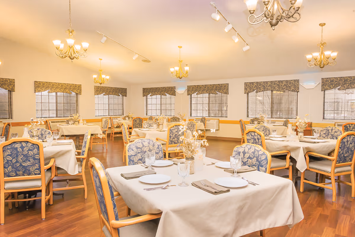 A spacious dining room with multiple tables covered in white tablecloths, each set with plates, glasses, napkins, and cutlery. The chairs have wooden frames with blue floral upholstery. The room has several windows with floral valances, wooden flooring, and multiple chandeliers and wall-mounted lights providing warm illumination.