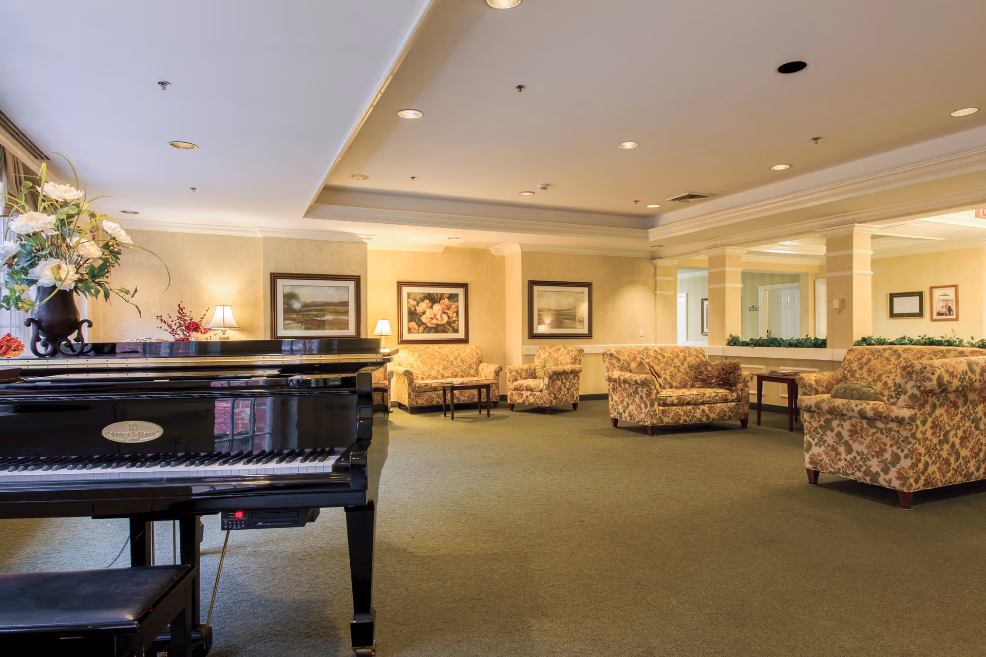 Spacious senior-living lounge featuring a grand piano, floral seating, and warm lamps.