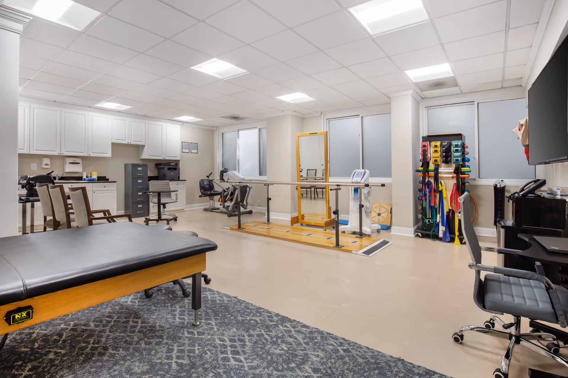A spacious physical therapy or rehabilitation room with exercise equipment including parallel bars, a large mirror, resistance bands, and chairs. There is a padded therapy table in the foreground and cabinetry with office equipment along the back wall. The room has a clean, bright appearance with overhead fluorescent lighting and frosted windows.