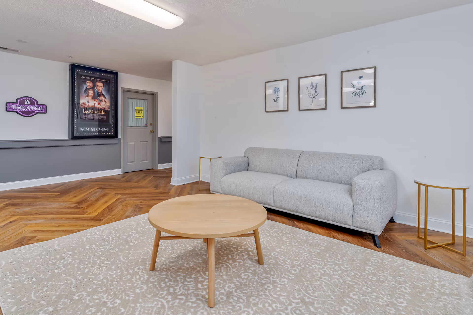 A senior living facility common area with a light gray sofa, a round wooden coffee table on a patterned beige rug, and two small side tables with gold legs and white tops. The walls are white with three framed botanical prints above the sofa. In the background, there is a gray door with a yellow sign and a wall-mounted theater sign and poster for Les Miserables.