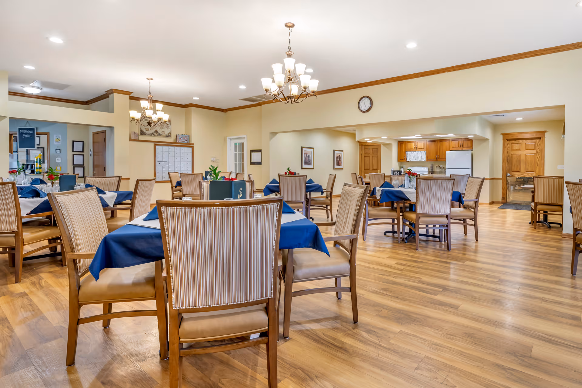 A spacious dining room with multiple tables covered in blue tablecloths and surrounded by wooden chairs with striped upholstery. The room has wooden flooring, cream-colored walls with wooden trim, and two chandeliers hanging from the ceiling. In the background, there is a kitchen area with wooden cabinets, a refrigerator, and a stove. A clock is mounted on the wall above the kitchen entrance.