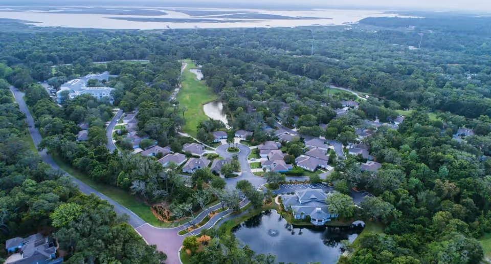 Aerial view of a senior living community with clustered single-story buildings, a central pond and fountain, winding roads, and dense surrounding trees.