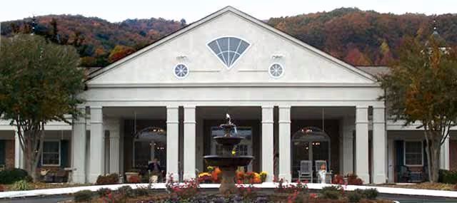 Front exterior view of a senior living facility building with white columns, a triangular pediment with decorative windows, a central black fountain surrounded by flowers, and trees on either side with hills in the background.
