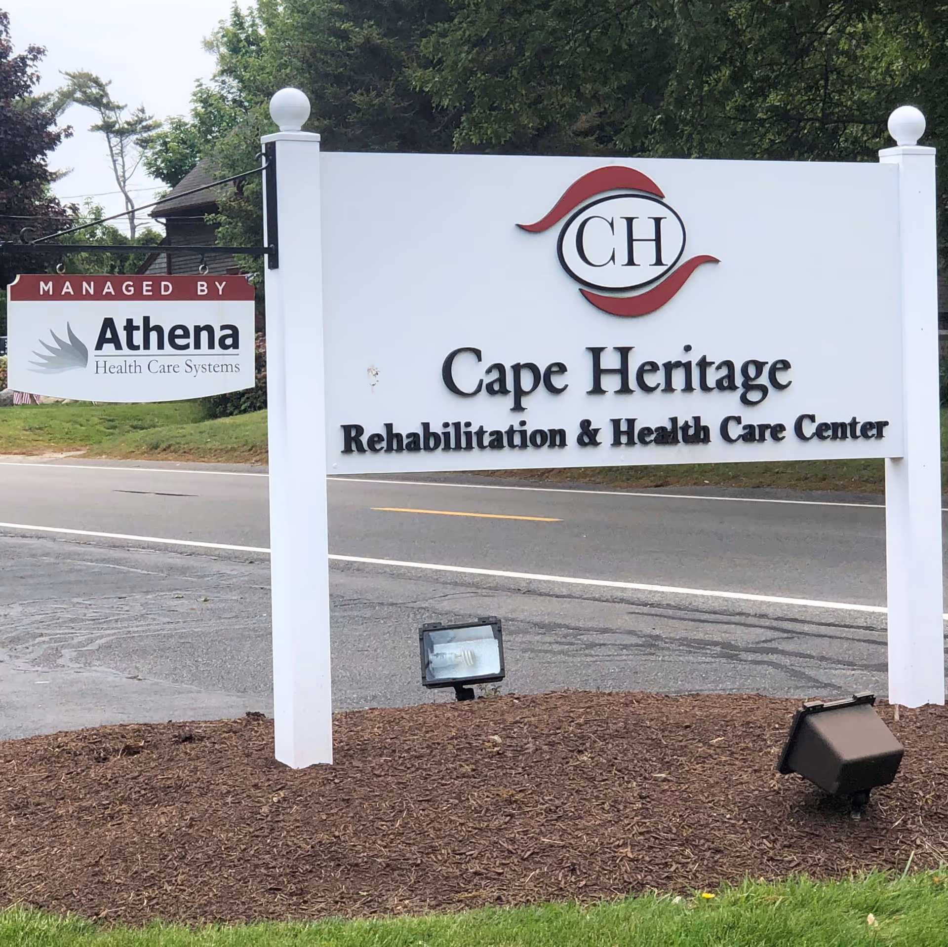 Outdoor sign for Cape Heritage Rehabilitation & Health Care Center with a smaller sign indicating it is managed by Athena Health Care Systems, located near a road with trees and grass in the background.