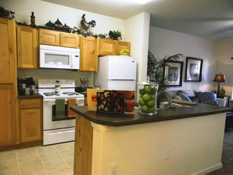 A kitchen area with wooden cabinets, a white microwave above a white stove, and a white refrigerator. There is a countertop with a sink and a glass jar filled with green apples. In the background, a living room area is partially visible with a couch, framed pictures on the wall, and a lamp.