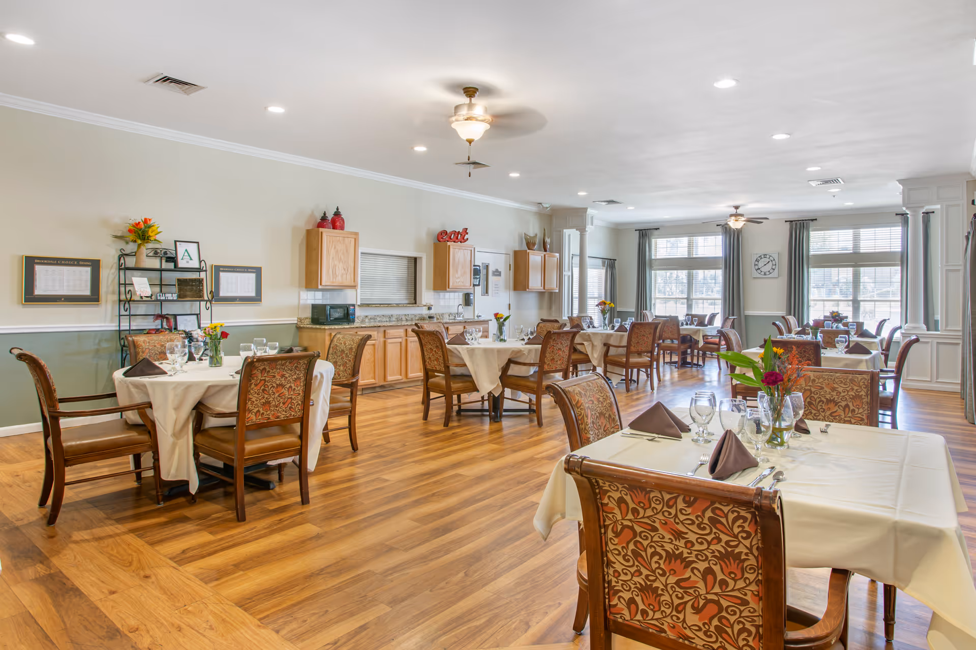 A bright and spacious dining room with multiple tables covered in white tablecloths, each set with glasses, silverware, and brown folded napkins. The chairs have patterned upholstery. The room features wooden flooring, large windows with curtains allowing natural light, ceiling fans, and a counter area with cabinets and a microwave. Decorative items and framed menus are on the walls.