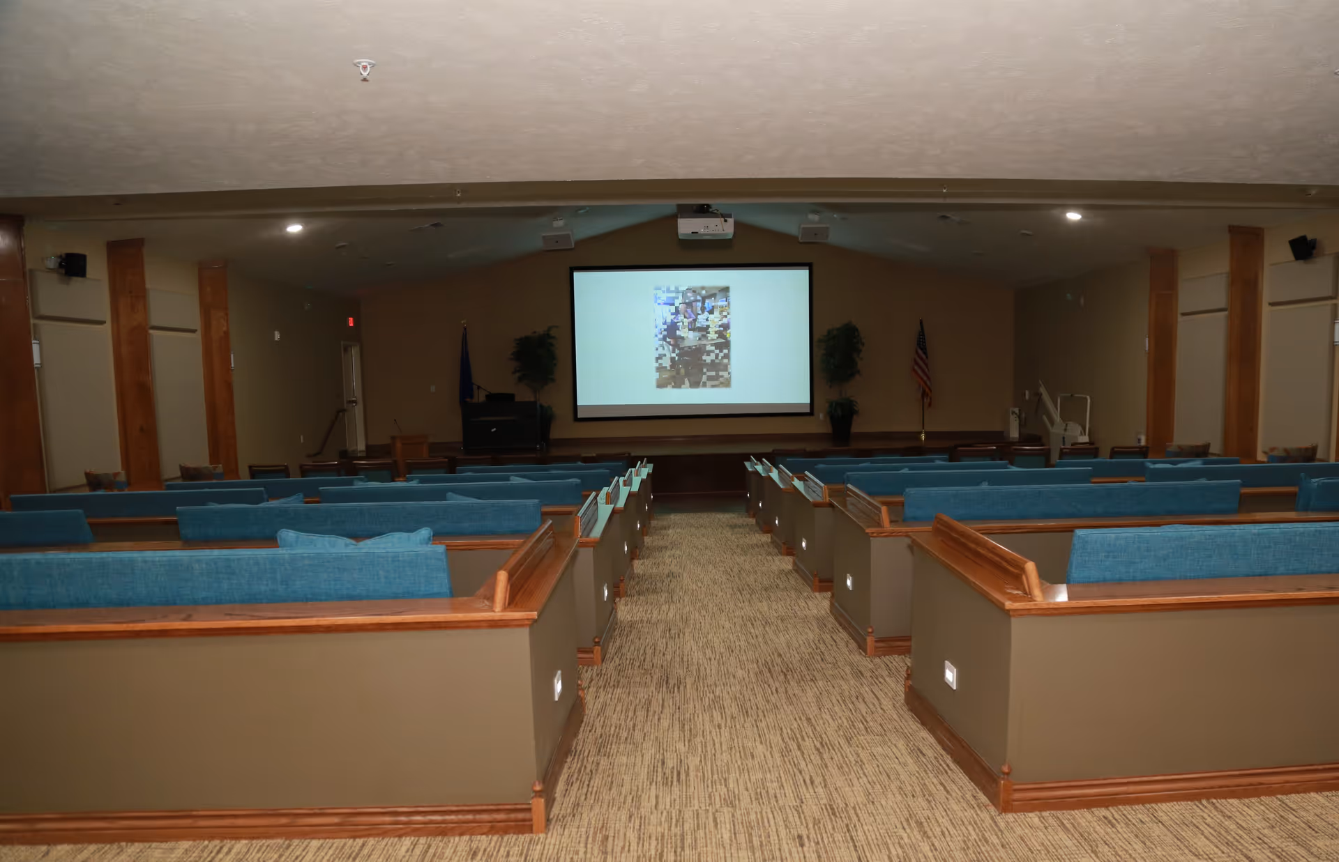 Interior view of a conference or presentation room with rows of blue cushioned pew-style seating facing a large projection screen at the front. The room has beige walls, carpeted floor, wooden trim, and two potted plants flanking the screen. An American flag is visible on the right side near the front.