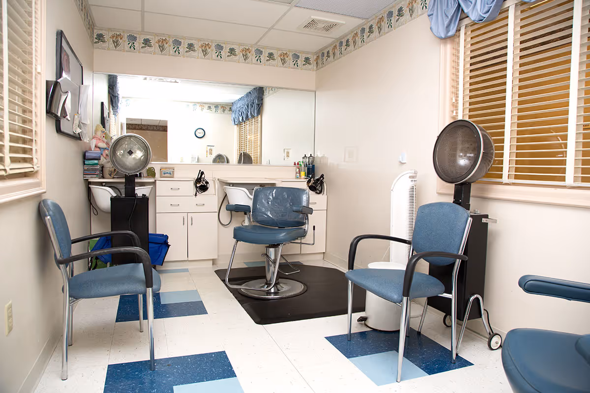 Interior of a small salon area with two blue salon chairs, two blue visitor chairs, a large mirror on the back wall, a counter with drawers, and two hair dryer stations. The floor has a white and blue checkered pattern, and there are windows with blinds and a blue valance.