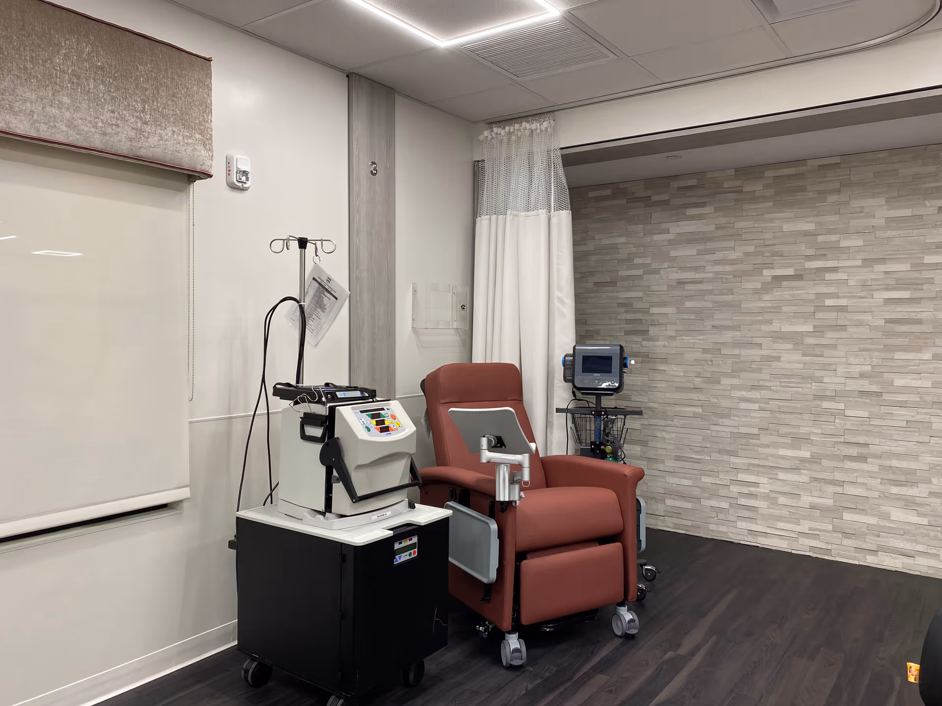 A medical treatment room with a red reclining chair on wheels, medical equipment on a black cart, an IV stand, and a monitor on a stand against a wall with light-colored textured tiles. The room has dark wood flooring and white walls with a window covered by a roller shade.