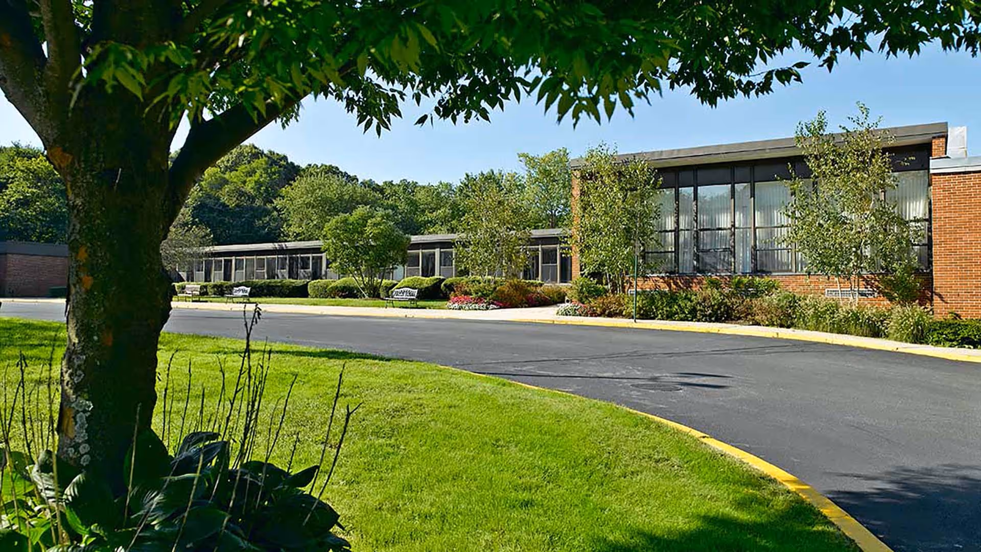 Exterior view of a single-story brick building with large windows, surrounded by well-maintained landscaping including trees, bushes, and a green lawn. A curved asphalt driveway with a yellow curb runs in front of the building under a clear blue sky.