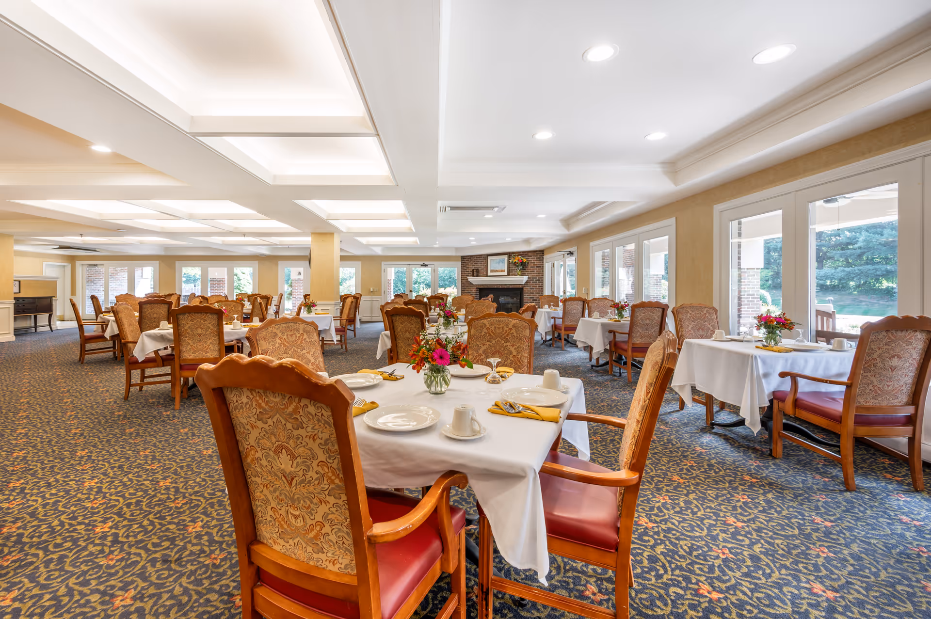 A spacious dining room with multiple tables covered in white tablecloths, each set with plates, cups, and yellow napkins. The chairs have wooden frames with patterned upholstery and red cushions. There are large windows and glass doors letting in natural light, and a fireplace with a painting and flowers on the mantel in the background. The carpet has a blue and gold floral pattern.
