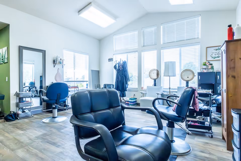 Bright and spacious salon area with multiple black salon chairs, large windows with blinds allowing natural light, a tall mirror, hair drying machines, and various salon equipment and storage units.
