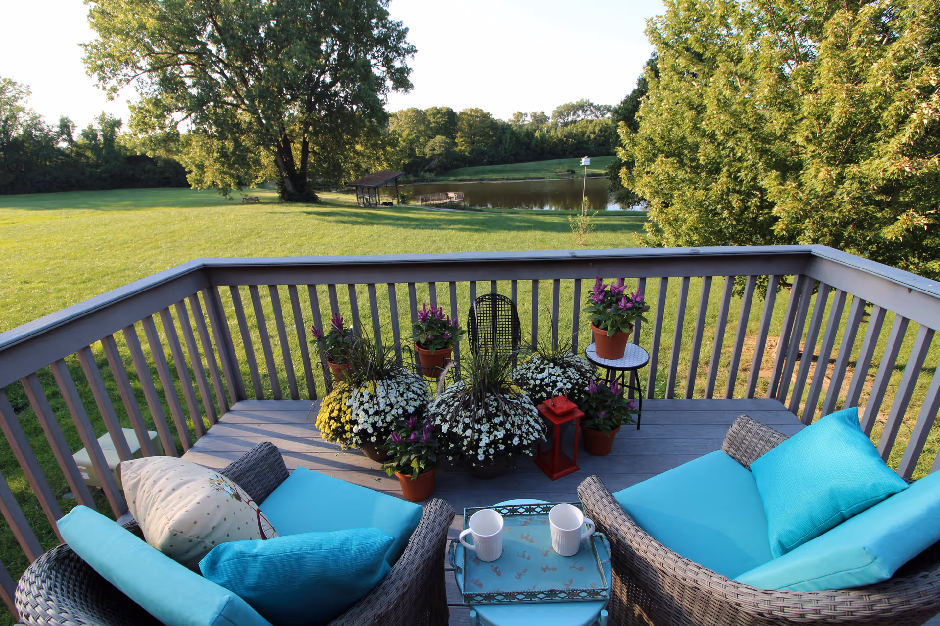 A cozy outdoor deck area with two wicker chairs featuring turquoise cushions and pillows, a small table with two white mugs, and several potted flowering plants. The deck overlooks a large grassy lawn with trees and a pond in the background under a clear sky.