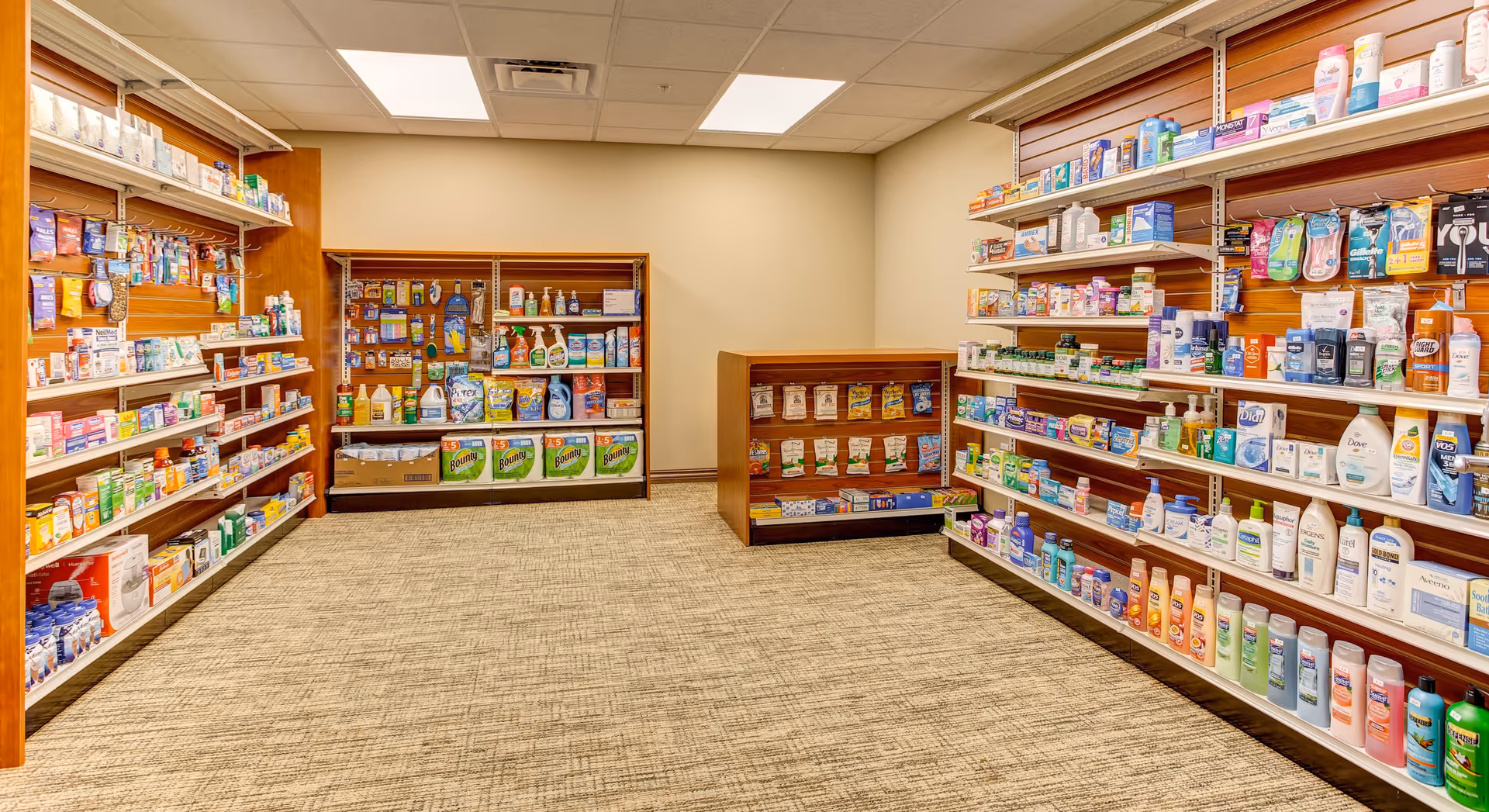 Interior view of a small retail store or convenience shop within a facility, featuring shelves stocked with various household and personal care products such as lotions, shampoos, cleaning supplies, and paper towels. The room has beige walls, a carpeted floor, and fluorescent ceiling lights.