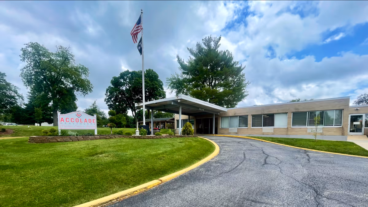 Exterior view of Accolade Healthcare Of Paxton Senior Living building with a driveway leading to a covered entrance. There is a green lawn with a sign displaying the facility's name, two flagpoles with the American flag and another flag, and trees in the background under a partly cloudy sky.