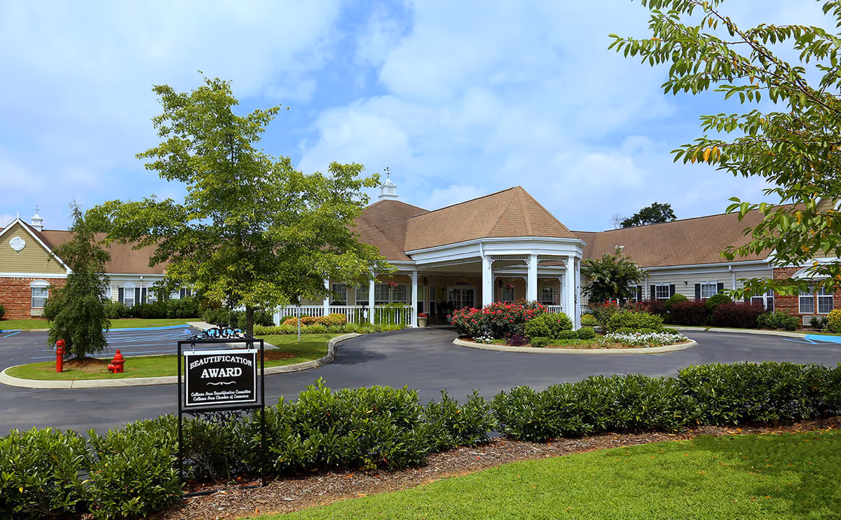 Exterior view of The Neighborhood at Cullman senior living facility showing a circular driveway with landscaped flower beds and bushes, a covered entrance with white columns, and a building with a brown roof and light-colored walls under a partly cloudy sky.