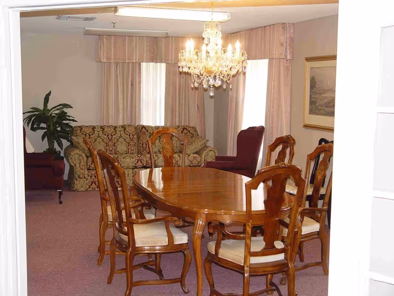 Wooden dining table with six chairs under a crystal chandelier in a common room with a sofa and armchairs.