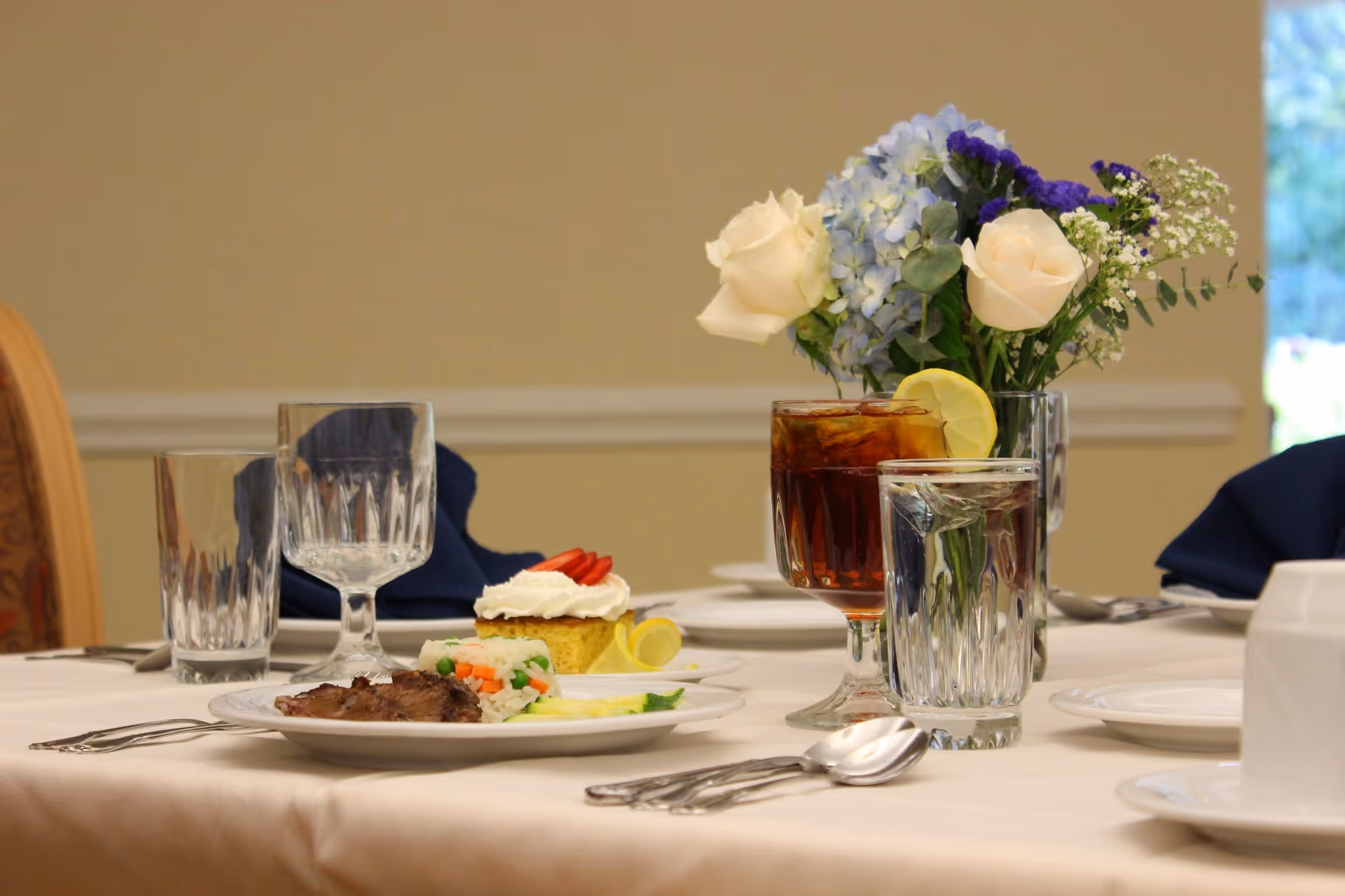 A dining table set with a plate of food including meat, rice with vegetables, and a dessert topped with whipped cream and strawberries. There are glasses of water and iced tea with a lemon slice, silverware, and a vase with white and blue flowers on the table.