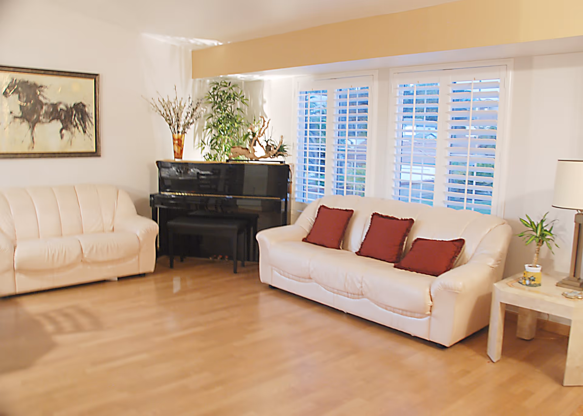 A bright living room with two cream-colored sofas, one with three red cushions, a black upright piano with a bench, a side table with a lamp and a small plant, and a large window with white shutters. There is a framed painting of a horse on the wall and decorative plants on and around the piano.