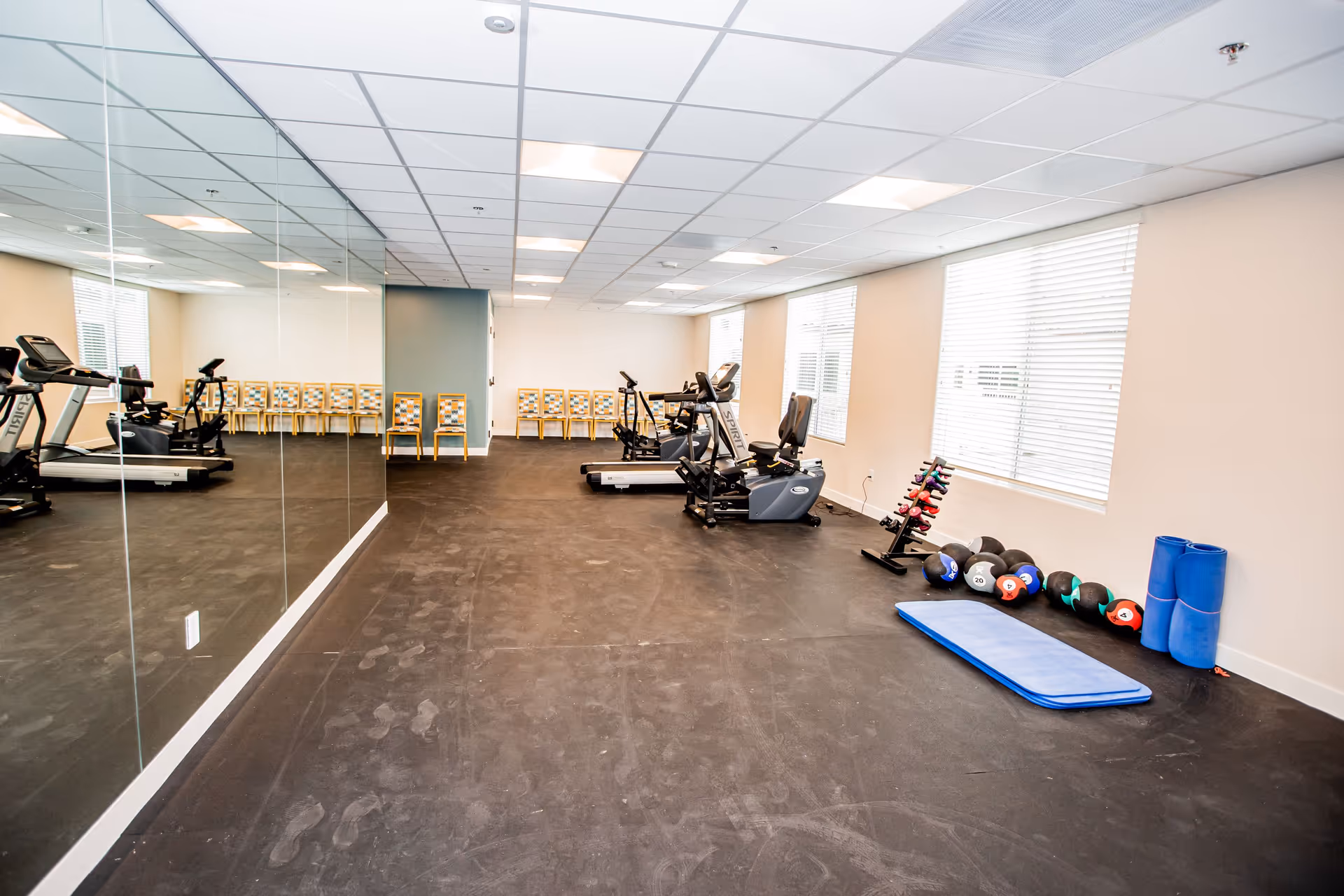 A fitness room with exercise equipment including treadmills, stationary bikes, a rack of dumbbells, medicine balls, and exercise mats. There is a large mirror on one wall and several chairs lined up against the far wall. The room has large windows with blinds and a black rubber floor.