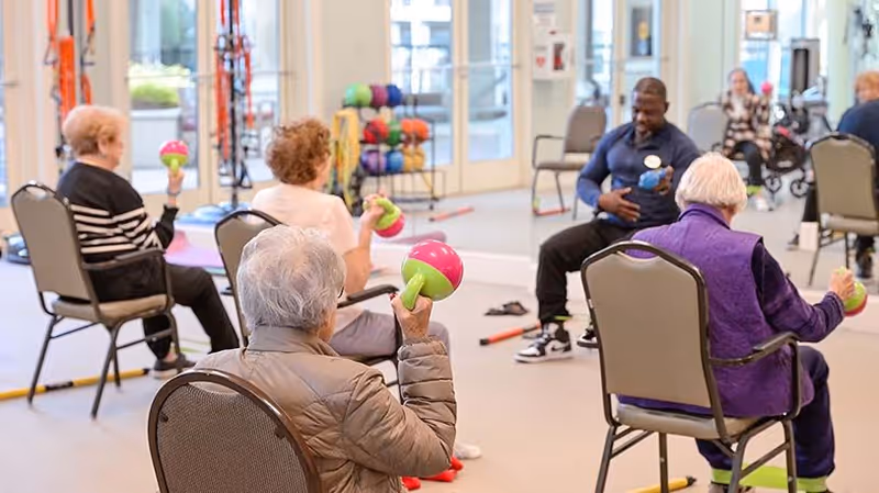 A group of elderly individuals seated in chairs participating in a fitness class inside a bright room. They are holding colorful hand weights and following the instructor, who is also seated and demonstrating exercises. The room has large windows and exercise equipment in the background.