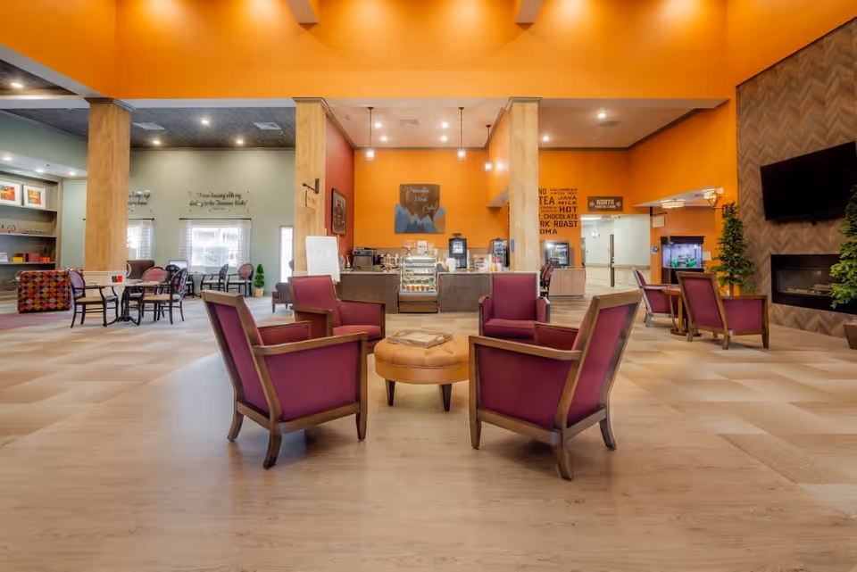 A spacious and brightly lit common area with four purple armchairs arranged around a round tan ottoman in the center. Behind the seating area is a coffee and snack bar with a sign that reads 'Princeton Drink Cafe'. The walls are painted in warm orange and neutral tones, and there is a fireplace with a mounted TV on the right side. Additional tables and chairs are visible in the background near large windows, and decorative plants are placed around the room.