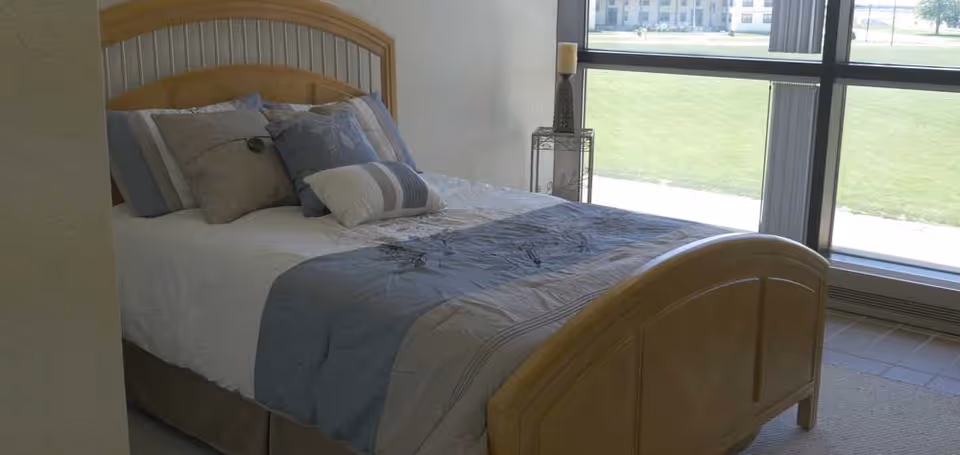 A bedroom with a wooden bed frame and a neatly made bed featuring a white and blue comforter and several pillows. The room has a large window with vertical blinds, allowing natural light to brighten the space and providing a view of a grassy outdoor area.