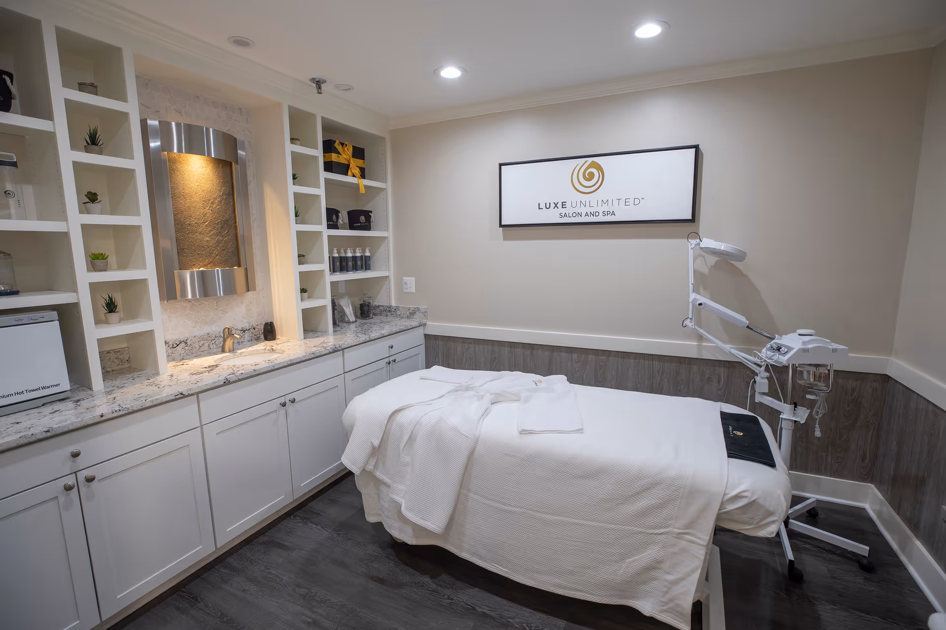 A spa treatment room with a massage table covered in white linens and towels. The room has white cabinets with a granite countertop, small potted plants on shelves, a wall-mounted water feature, and a sign that reads 'Luxe Unlimited Salon and Spa'. There is also a piece of spa equipment with an adjustable arm next to the table.