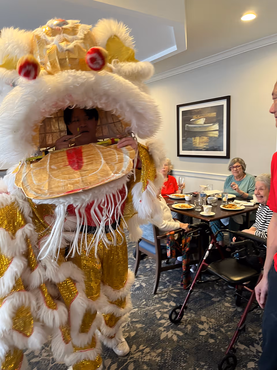 A person dressed in a traditional gold and white lion dance costume performing indoors near a dining area where elderly people are seated at a table with plates of food, smiling and watching the performance.