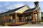Exterior view of a single-story building with a covered entrance supported by wooden pillars, a wheelchair accessible ramp, and a small bell tower on the roof. The building has brick and siding walls, with a landscaped area featuring flowers in front.