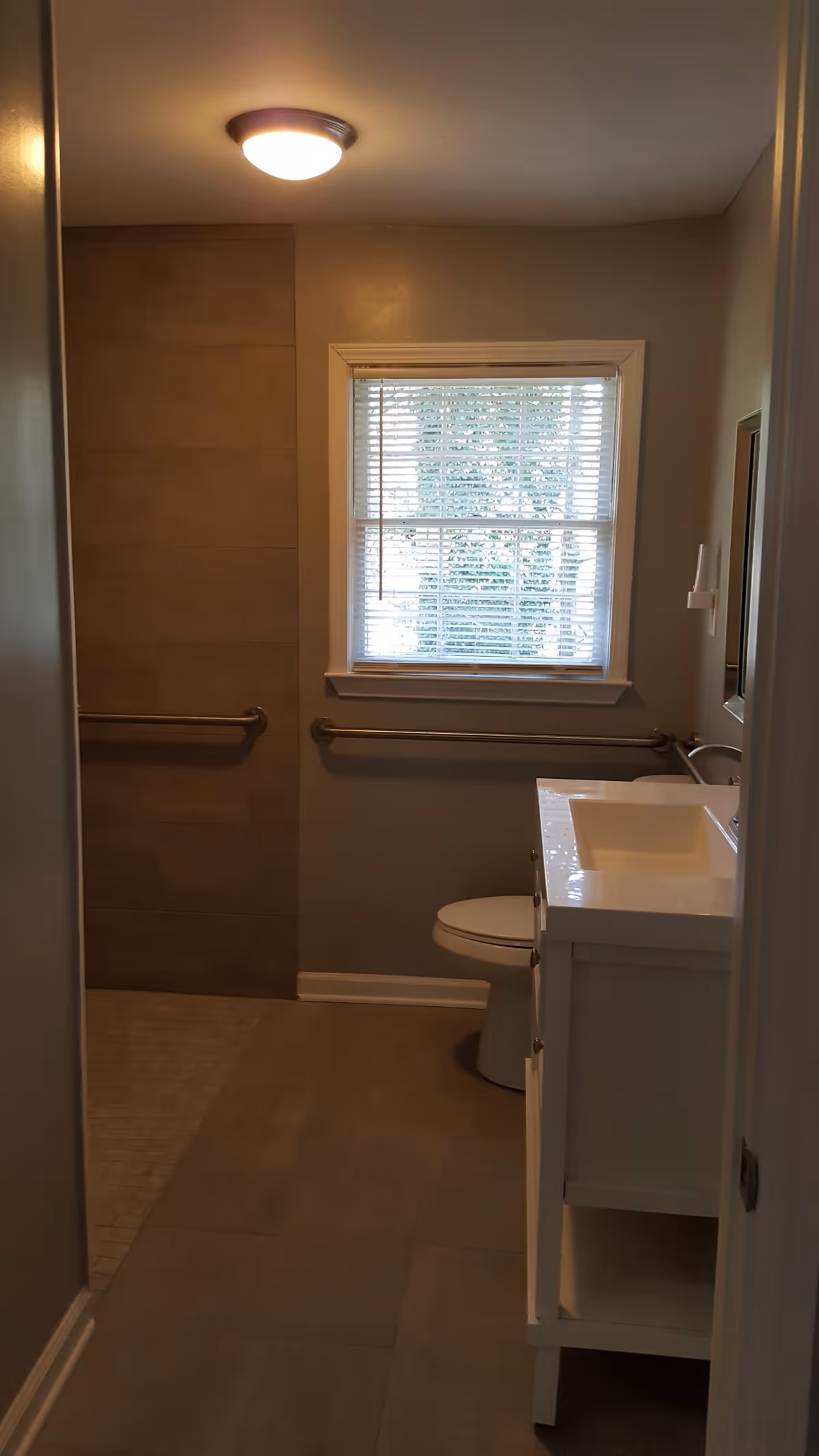 A bathroom with a window covered by white blinds, a white vanity with a sink, a toilet, and grab bars installed on the walls near the toilet and in the shower area. The floor and shower walls are tiled, and there is a ceiling light fixture.