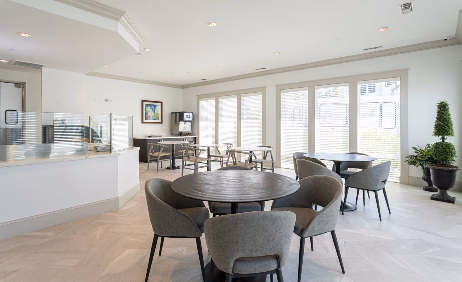 A bright dining area with round tables and gray upholstered chairs. Large windows with blinds allow natural light to fill the room. There is a counter with a glass partition on the left side and an ice machine against the far wall. A framed picture hangs on the wall, and there are decorative plants near the windows.