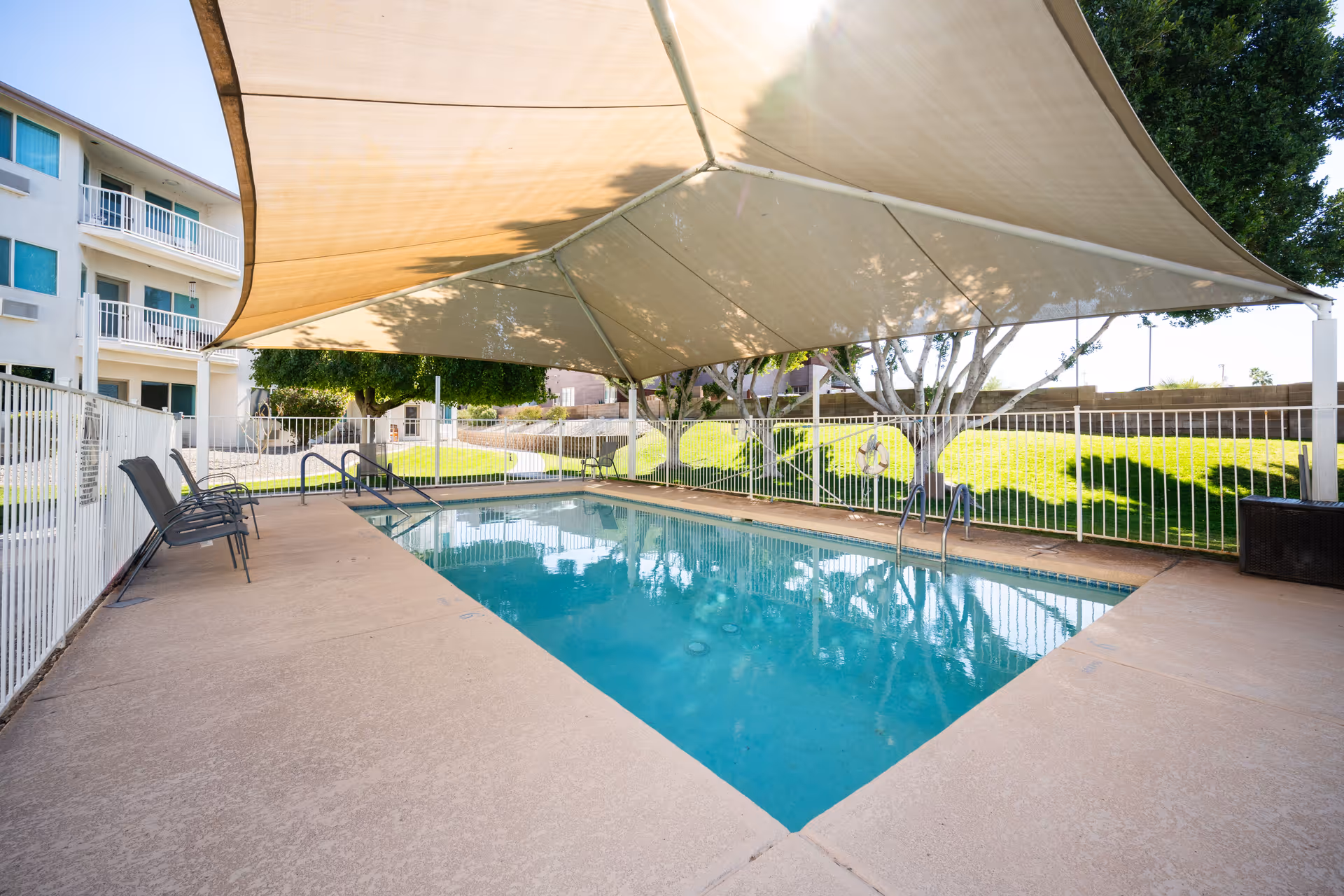 Outdoor swimming pool area with a large beige shade canopy overhead, surrounded by a white safety fence. Several chairs are placed along the poolside. In the background, there is a multi-story building with balconies and green grass with trees.