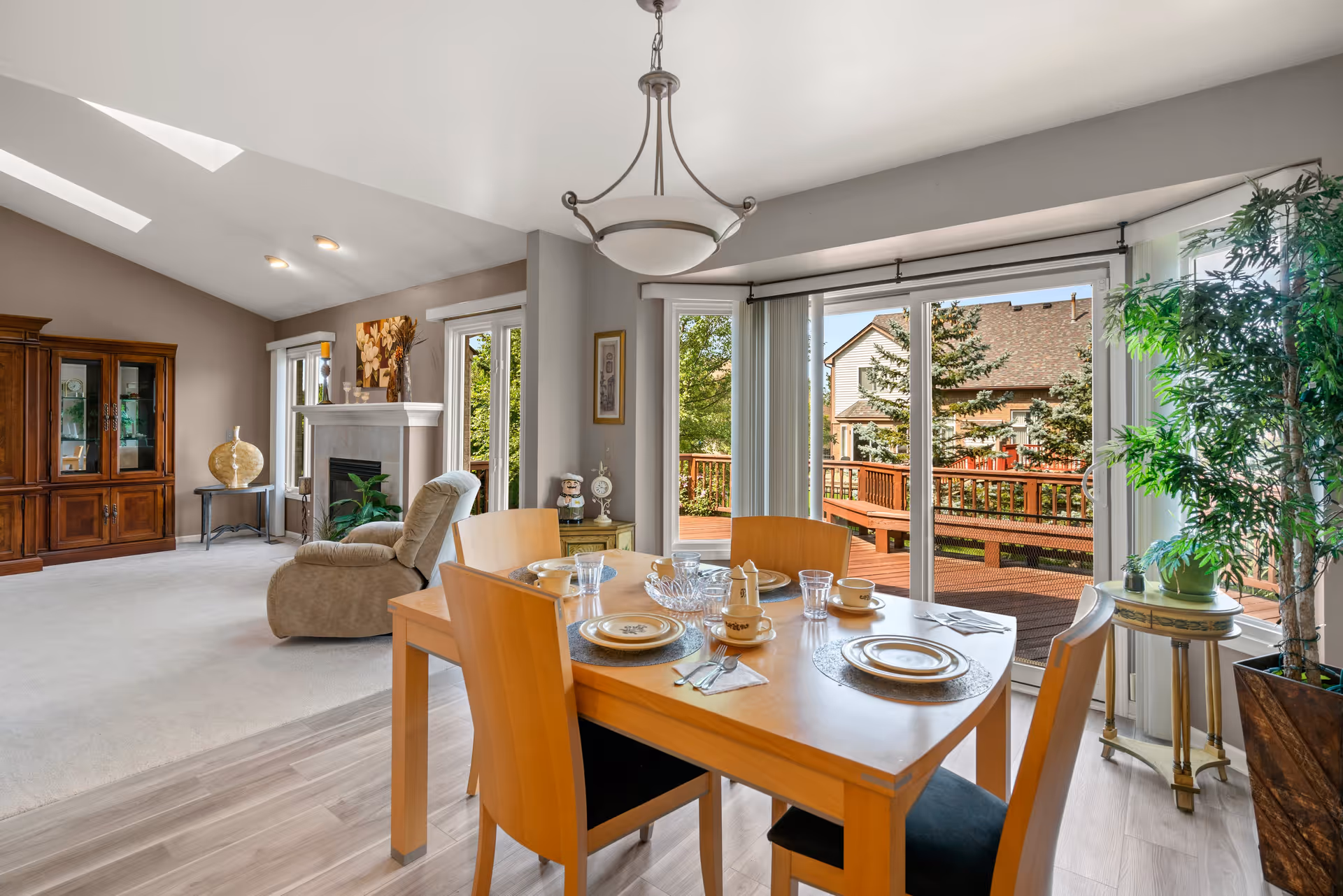 A bright and spacious living and dining area with a wooden dining table set for four, large sliding glass doors leading to an outdoor deck, a comfortable recliner chair, a fireplace, and a wooden cabinet. The room features light-colored walls, a high ceiling with skylights, and a large potted plant near the window.