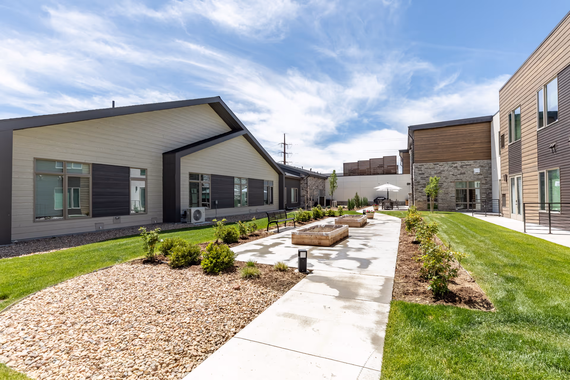 Outdoor courtyard area at Terra Bluffs featuring a concrete walkway flanked by landscaped garden beds with small shrubs and grass. Modern buildings with large windows surround the courtyard under a partly cloudy blue sky. Benches and raised garden beds are visible along the walkway, with a seating area and umbrella in the background.