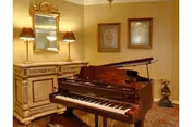 A warmly lit room featuring a polished grand piano in the foreground. Behind the piano is a decorative wooden sideboard with two lamps and a large mirror mounted on the wall. Two framed artworks hang on the adjacent wall, and a small decorative plant is placed on the floor to the right.