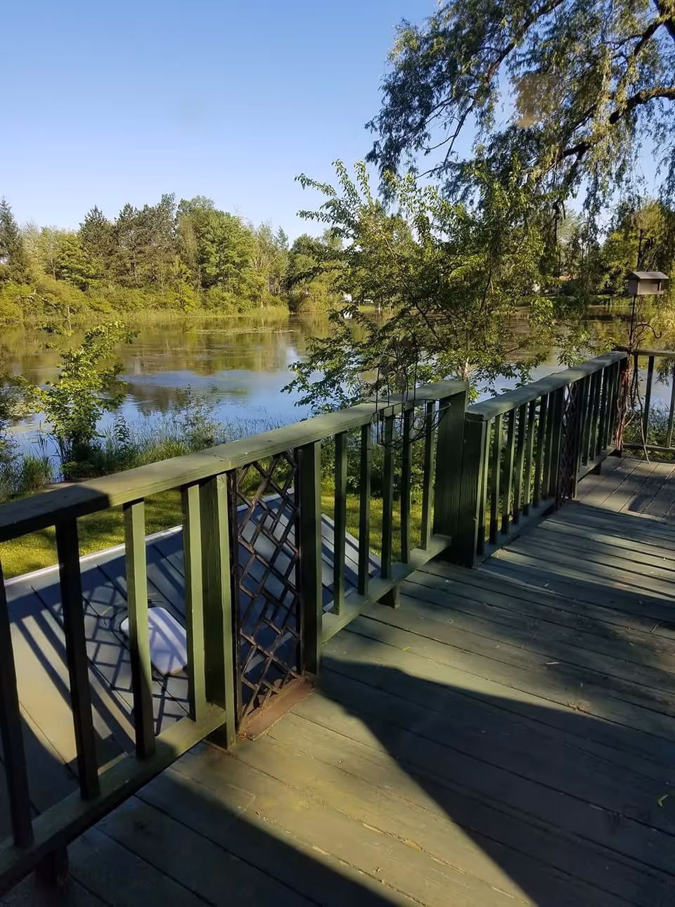 View from a wooden deck with green railings overlooking a calm pond surrounded by trees and greenery under a clear blue sky.