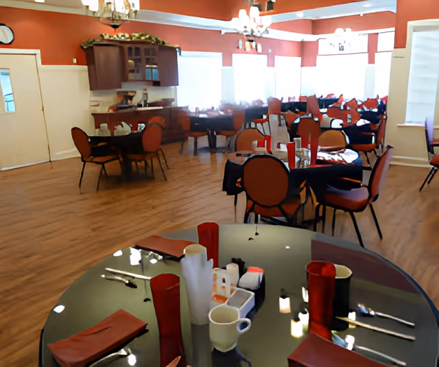 Interior view of a dining room with multiple round tables set with red napkins, cups, and utensils. The room has wooden flooring, orange walls, large windows letting in natural light, and chandeliers hanging from the ceiling. There is a cabinet with glass doors and decorative greenery on top against one wall.