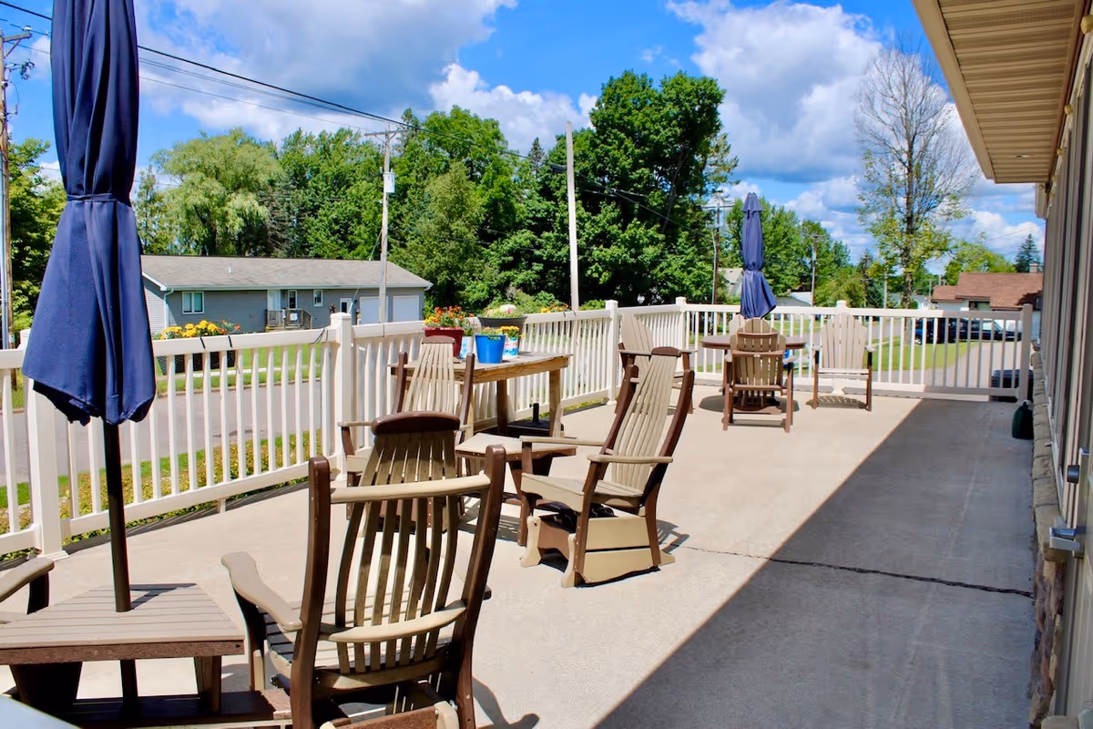 Outdoor patio area with several wooden chairs and tables, some with closed blue umbrellas. The patio is surrounded by a white railing and overlooks a street with houses and green trees under a partly cloudy blue sky.