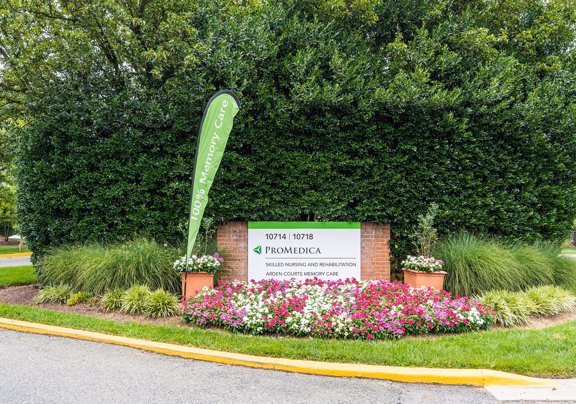 Outdoor view of a Promedica sign for Arden Courts Memory Care, surrounded by green bushes, grass, and colorful flowers. A green flag next to the sign reads '100% Memory Care'.