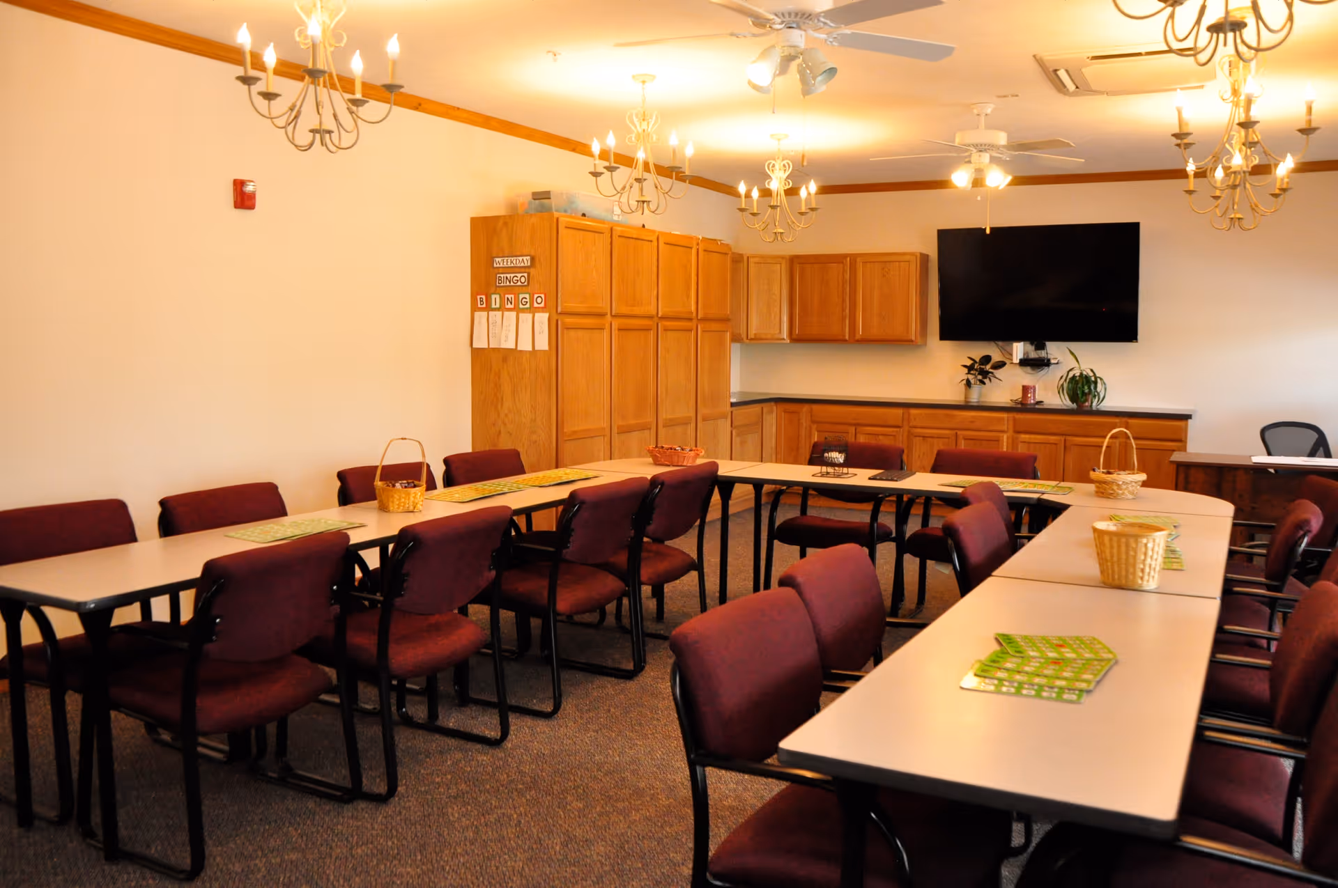 A well-lit activity room with tables arranged in a U-shape, surrounded by maroon chairs. There are bingo cards and small baskets on the tables. The room has wooden cabinets along one wall, a mounted flat-screen TV, ceiling fans, and chandeliers providing light.